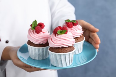 Pastry chef holding plate with sweet cupcakes on blue background, closeup