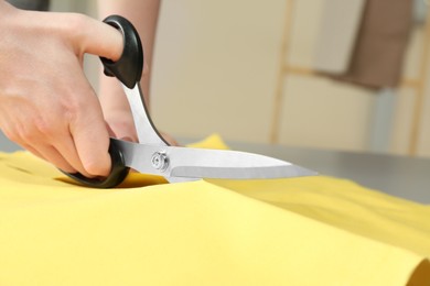 Photo of Seamstress cutting yellow fabric with scissors at workplace, closeup