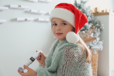 Photo of Cute little girl in Santa hat holding gift from New Year advent calendar at home