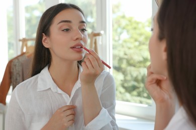 Beautiful young woman applying cosmetic pencil on lips near mirror indoors