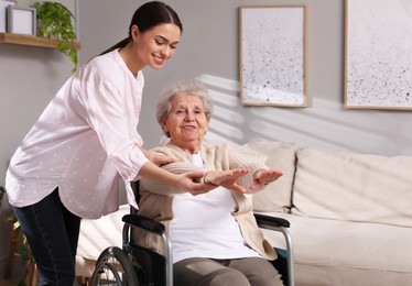 Senior woman in wheelchair doing physical exercise and young caregiver helping her indoors. Home health care service