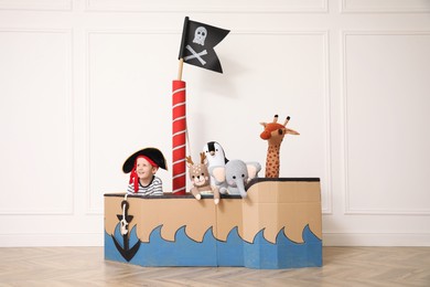 Photo of Cute little boy playing with toys in pirate cardboard ship near white wall indoors