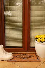 Door mat with word Welcome, stylish boots and beautiful flowers on floor near entrance