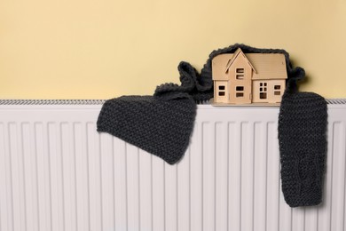 Wooden house model and knitted scarf on heating radiator near beige wall. Energy efficiency concept