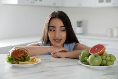 Woman choosing between fruits and burger with French fries in kitchen