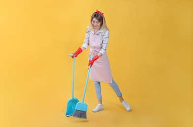 Young housewife with broom and dustpan on yellow background
