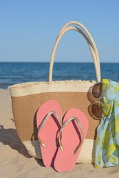 Straw bag with beach wrap, sunglasses and flip flops on sandy seashore. Summer accessories