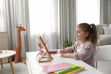 Adorable little girl doing homework with tablet at table indoors