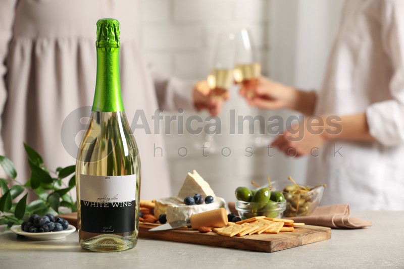 Women clinking glasses indoors, focus on table with bottle of wine and different snacks. Space for text