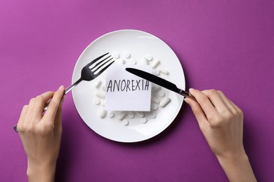 Woman holding cutlery over plate with pills and word Anorexia on purple background, top view