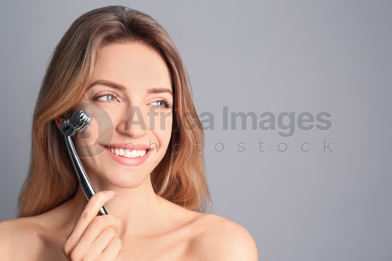 Photo of Young woman using metal face roller on light grey background, space for text