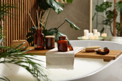 Wooden bath tray with open book, candle and body care products on tub indoors. Relaxing atmosphere