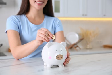 Young woman putting coin into piggy bank at table indoors, closeup. Money savings