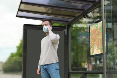 Young man in protective mask talking on phone while waiting for public transport at bus stop