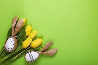 Photo of Easter bunnies made of craft paper and eggs among beautiful tulips on light green background, flat lay. Space for text