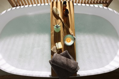 Wooden bath tray with candle and bathroom amenities on tub indoors, top view