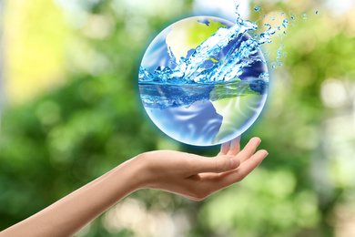 Woman holding icon of Earth with water splashes on blurred green background, closeup. Ecology concept