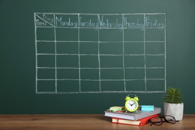 Alarm clock, stationery and plant on wooden table near green chalkboard with drawn school timetable