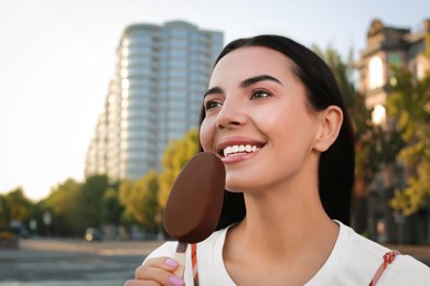 Beautiful young woman eating ice cream glazed in chocolate on city street