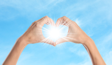 Woman making heart around bright light with hands, closeup. Solar energy concept