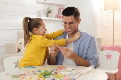 Happy father with his cute daughter making beaded jewelry at table in room