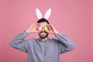 Photo of Happy man in bunny ears headband holding painted Easter eggs near his eyes on pink background