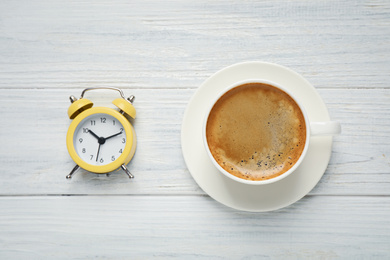 Cup of morning coffee and alarm clock on white wooden table, flat lay