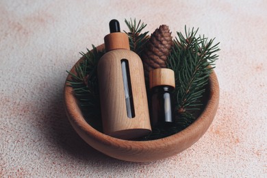 Bowl with bottles of aromatic essential oil, pine branches and cone on color table, closeup