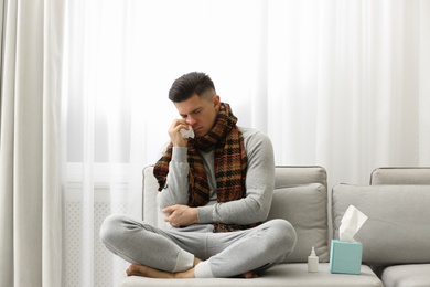 Ill man sitting near nasal spray and box of paper tissues on sofa at home