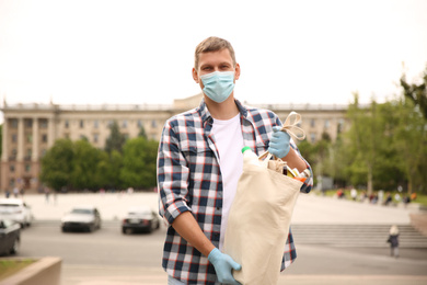 Male volunteer in protective mask and gloves with products on city street. Aid during coronavirus quarantine