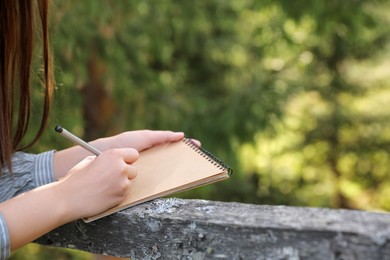 Young woman drawing with pencil in notepad near wooden fence, closeup