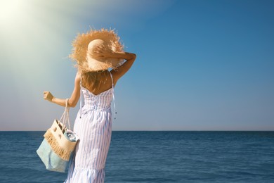 Woman with beach bag and straw hat near sea, back view. Space for text