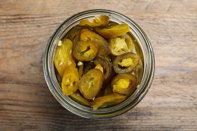 Photo of Glass jar with slices of pickled green jalapeno peppers on wooden table, top view