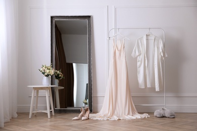 Elegant wedding dress and silk robe hanging on rack in room