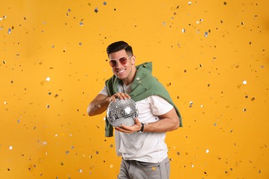 Happy man with disco ball and confetti on yellow background