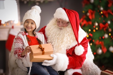 Little girl with gift box sitting on authentic Santa Claus' lap indoors