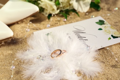 Gold wedding rings, invitation and decor on sandy beach, closeup