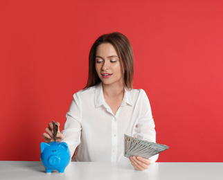 Young woman putting money into piggy bank at table on crimson background