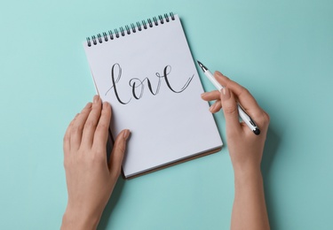 Woman writing word LOVE in notepad on turquoise background, top view