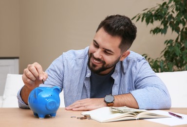 Young man putting coin into piggy bank at table indoors. Money savings