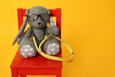 Toy bunny with stethoscope and glasses on chair against yellow background, space for text. Pediatrician practice