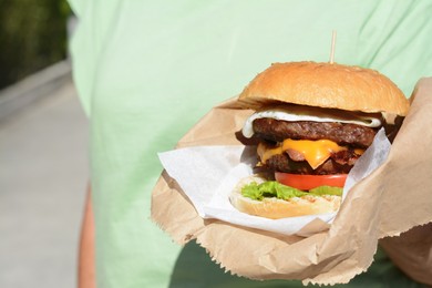 Photo of Woman holding delicious burger in paper wrap outdoors, closeup. Space for text