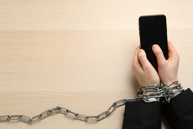 Man holding smartphone in chained hands at wooden table, top view. Internet addiction