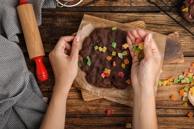 Woman adding candied fruits to unbaked chocolate cookie at wooden table, top view