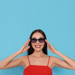 Attractive happy woman touching fashionable sunglasses against light blue background