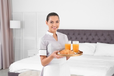 Young chambermaid holding tray with breakfast in bedroom