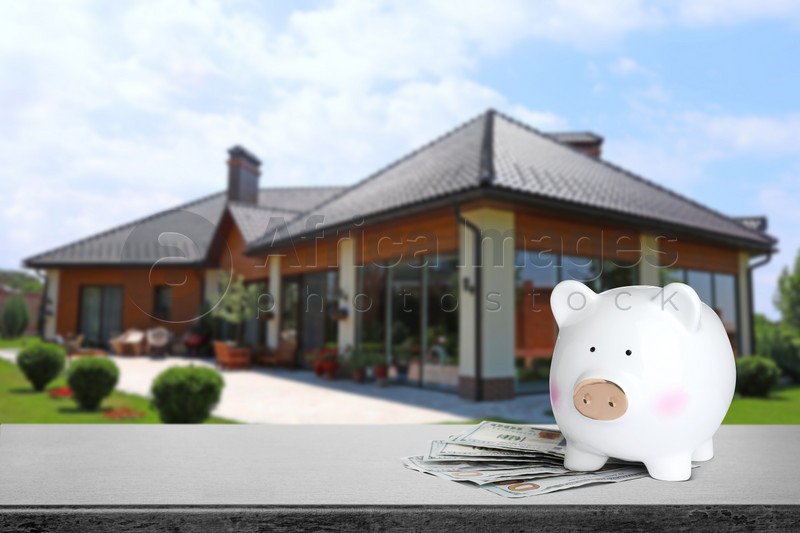 Piggy bank and money on stone surface and blurred view of beautiful house, space for text. Mortgage concept