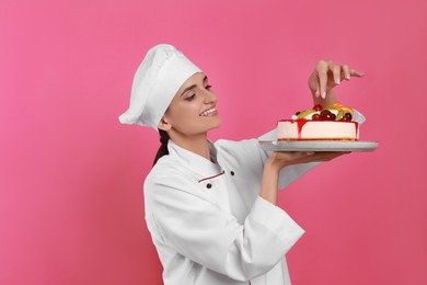 Happy professional confectioner in uniform decorating delicious cake on pink background