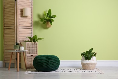 Stylish room interior with comfortable knitted pouf and plants near light green wall