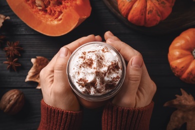 Woman holding tasty pumpkin latte at black wooden table, top view